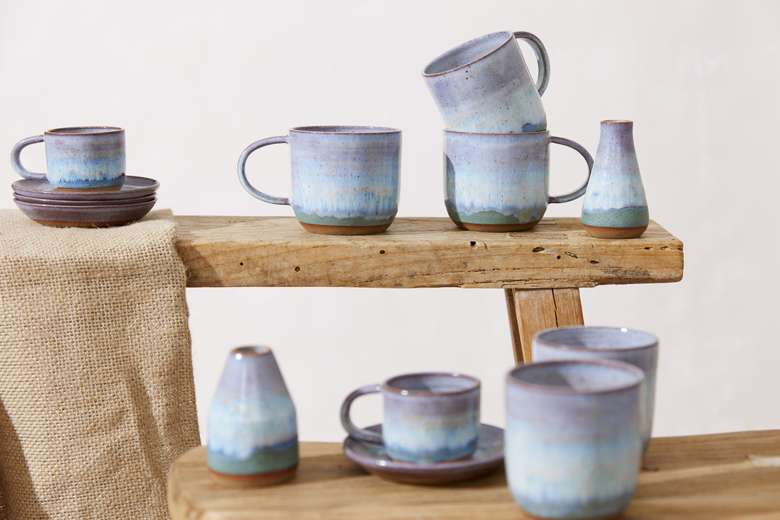 Handmade ceramic mugs, vases and plates in a purple, lavender, white and blue glaze. The handmade ceramics are in a coastal decor style and sitting on a wooden stool with a brown hessian mat.