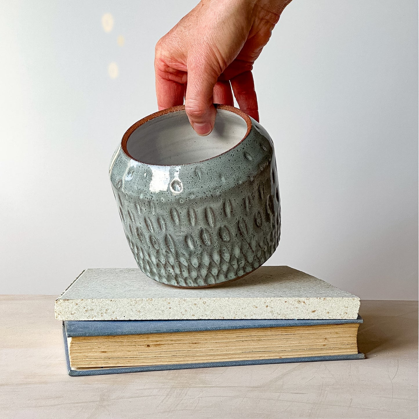 Person holding a textured ceramic pot over a stack of books on a light surface.