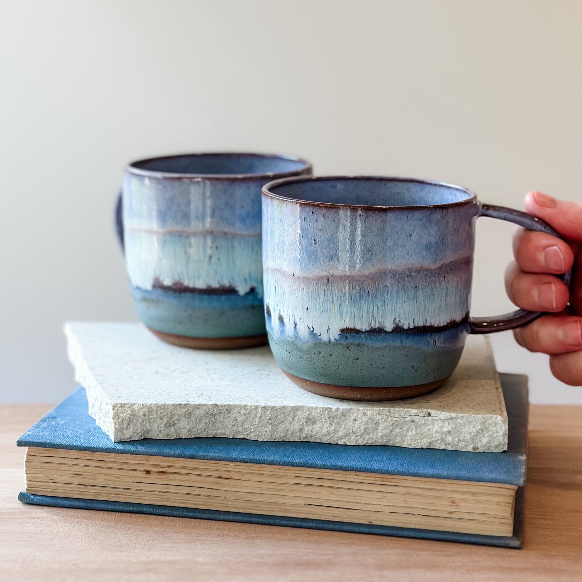 Two purple and blue ceramic mugs on top of books, with a hand holding one mug.