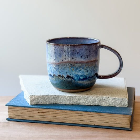 Purple and blue ceramic mug on top of a book and a tile with a plain background