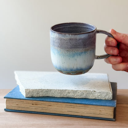 Hand holding a ceramic mug over a tile and a book on a wooden surface.