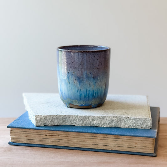 A purple and blue ceramic cup on top of a tile and a book with a plain background