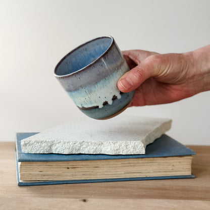 Hand holding a ceramic cup over a stack of books on a wooden surface.