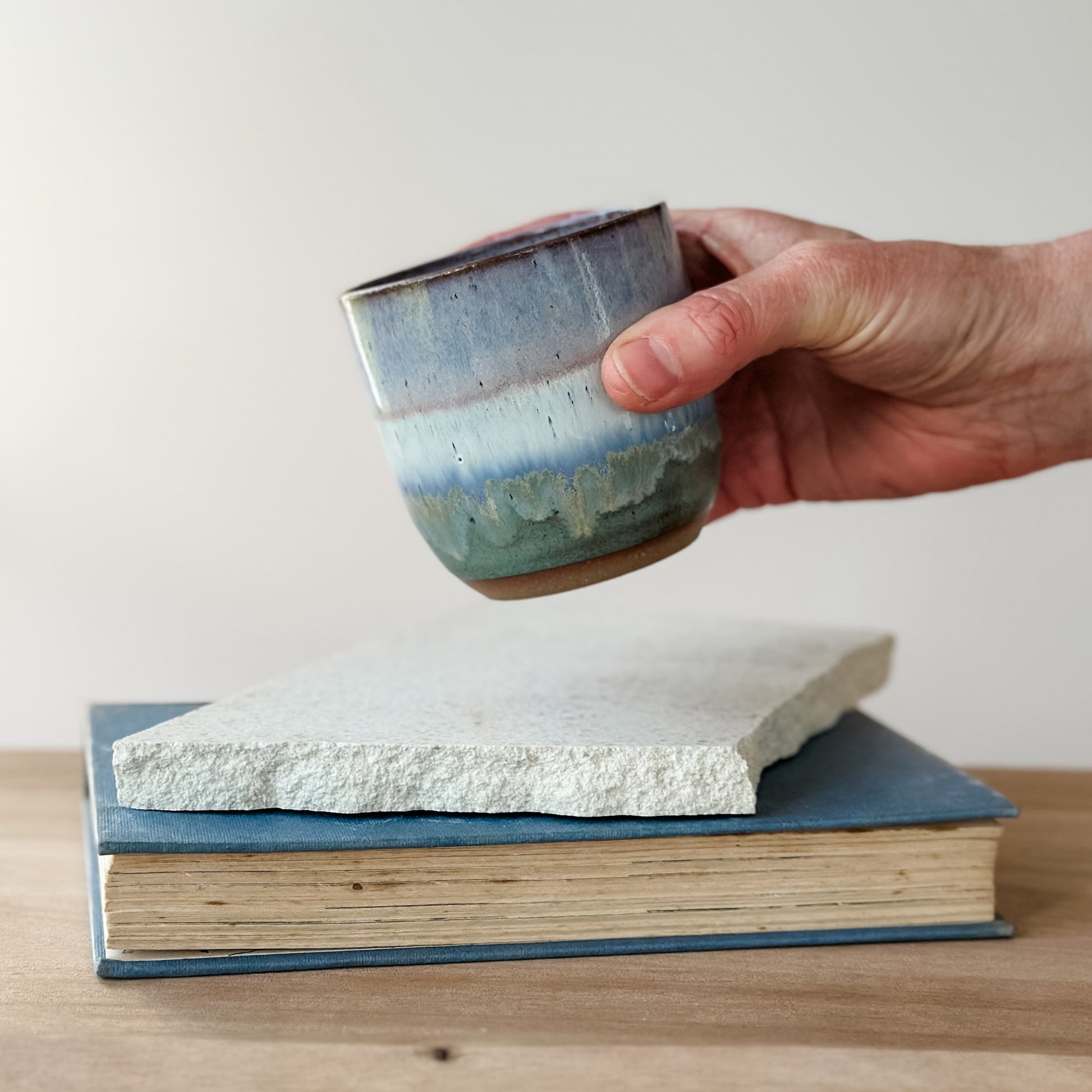Hand holding a ceramic cup over a stack of books on a wooden surface.