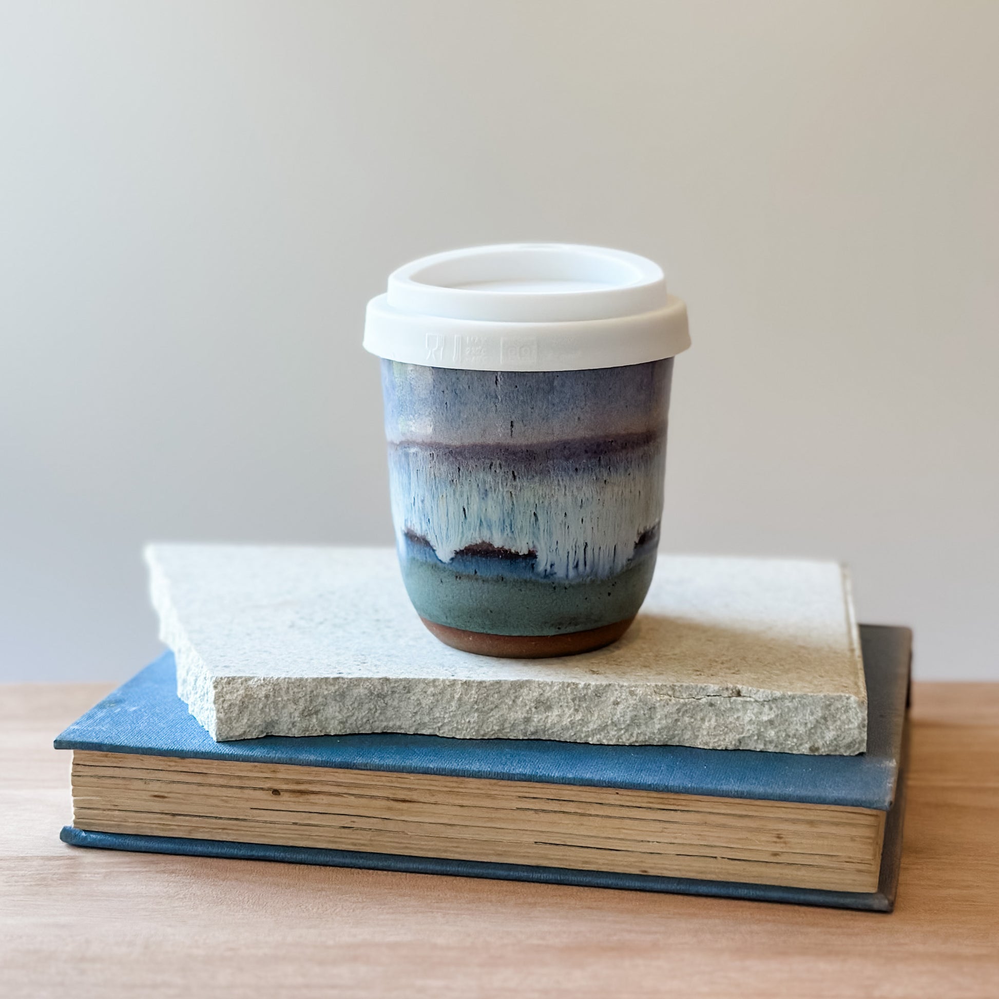 Ceramic cup with a white lid on top of a tile and a book on a wooden surface.