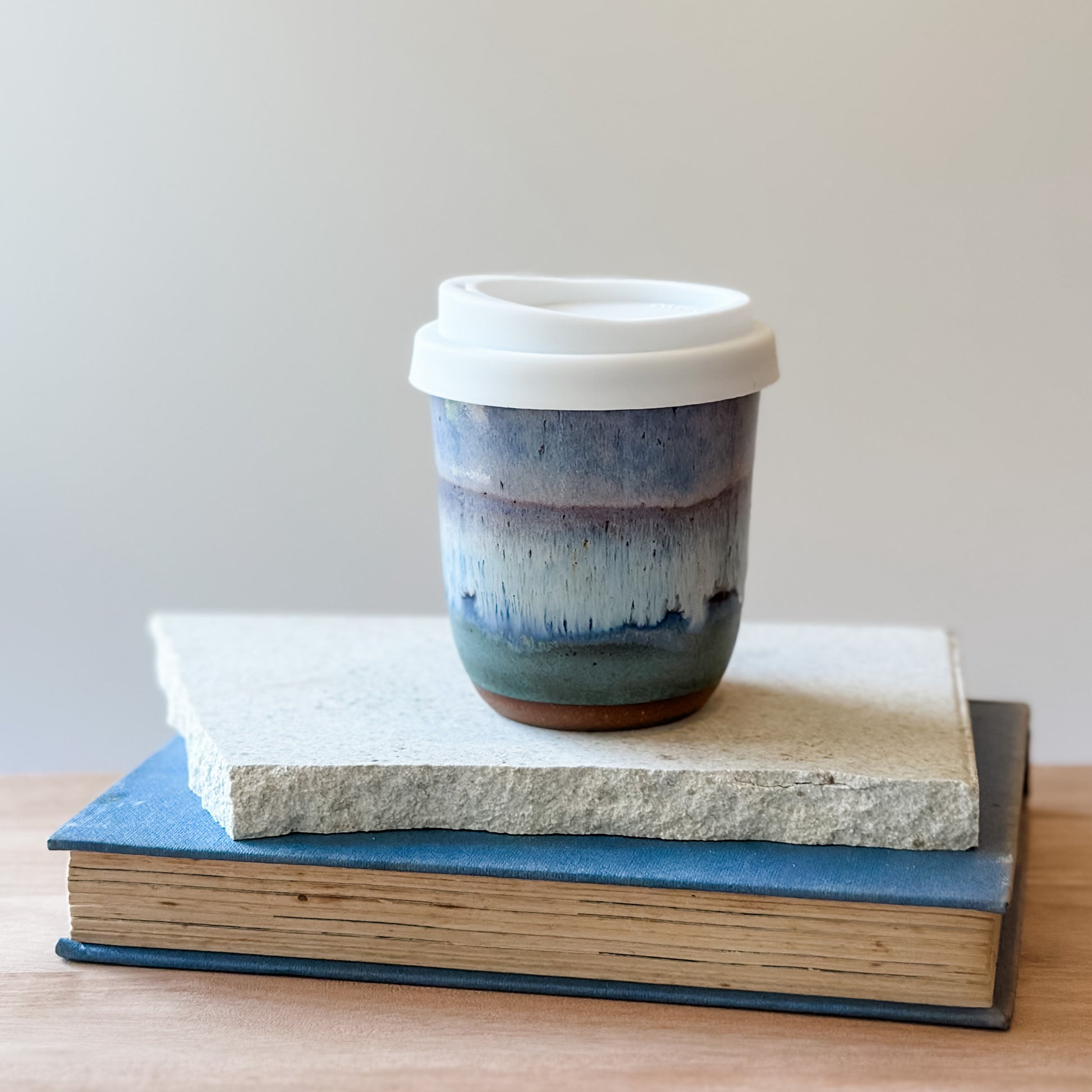 Ceramic travel mug with multicoloured glaze on top of a tile and a book