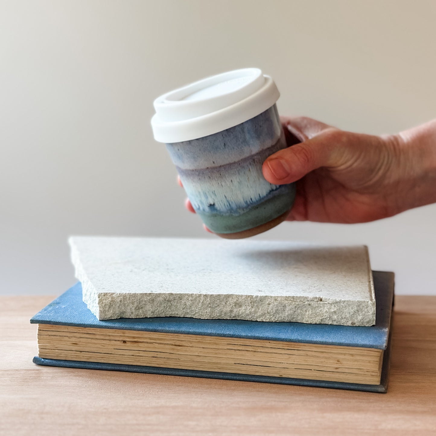 Hand holding a ceramic cup with a white lid over two stacked books on a wooden surface.