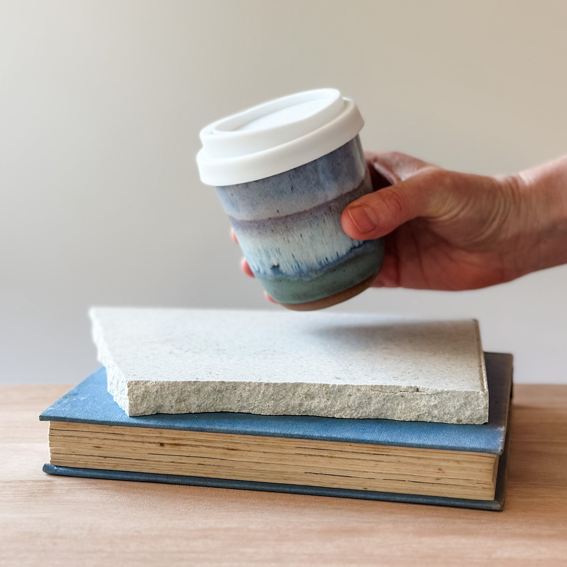 Hand holding a ceramic cup with a white lid over two stacked books on a wooden surface.