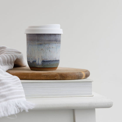 A handmade ceramic keepcup with a lavender, blue and white glaze. The handmade keep cup is sitting on a wooden board on top of a stool in a coastal setting.