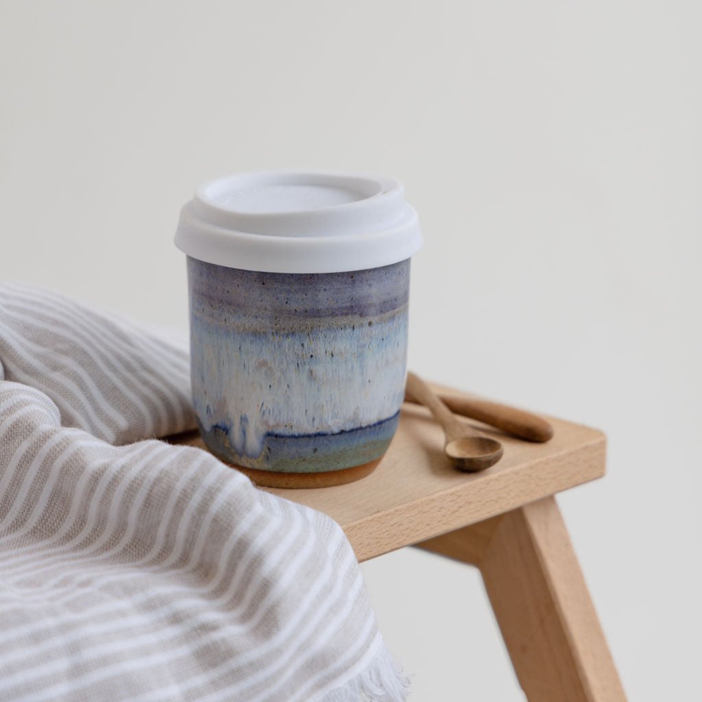 A top view of  the coastal handmade ceramic keepcup with a lavender, blue and white glaze. The handmade keep cup is sitting on a wooden board on top of a stool in a coastal setting.