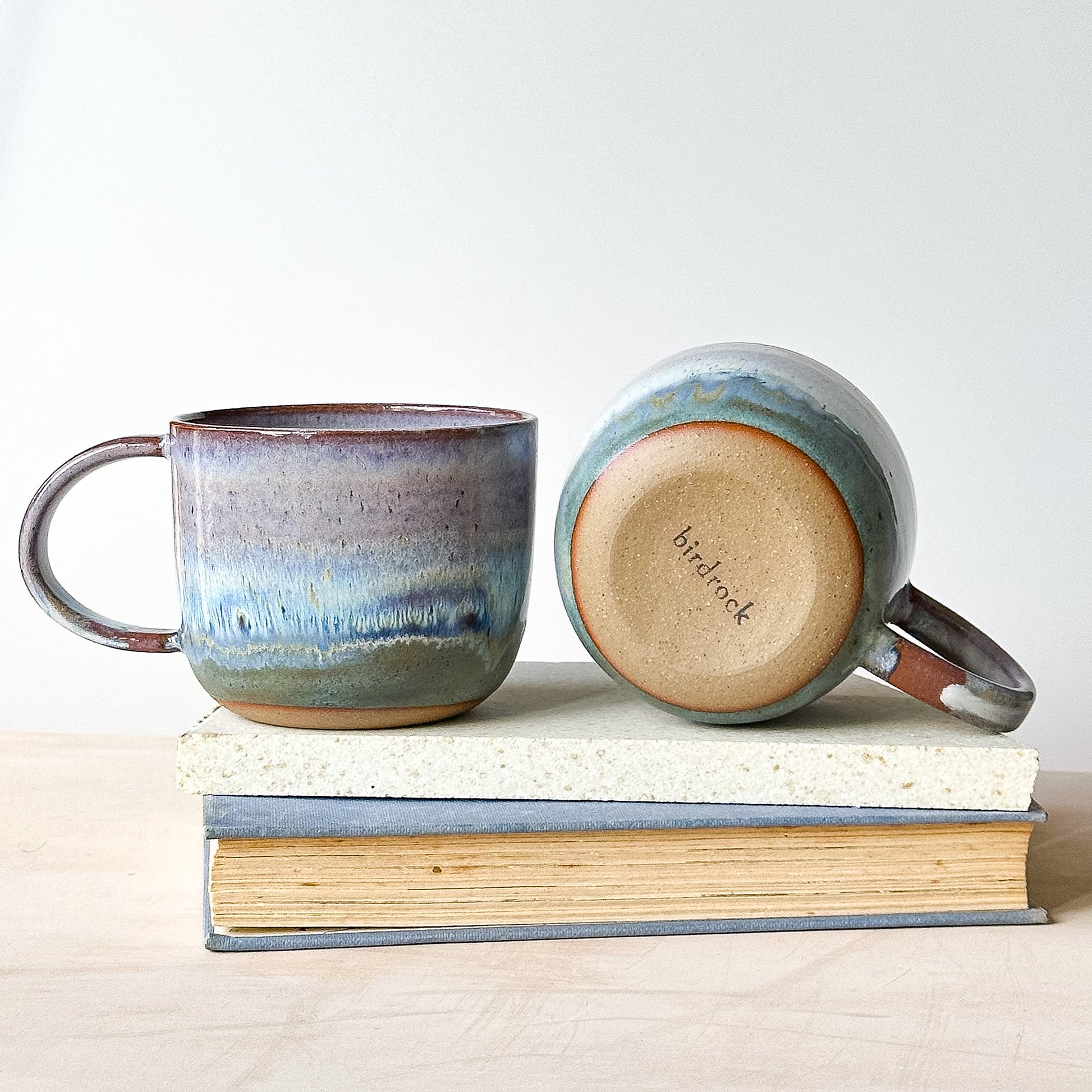 Two ceramic mugs with a gradient design on a stack of books against a white background