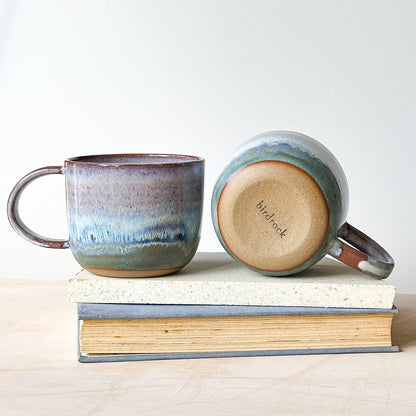 Two ceramic mugs with a gradient design on a stack of books against a white background