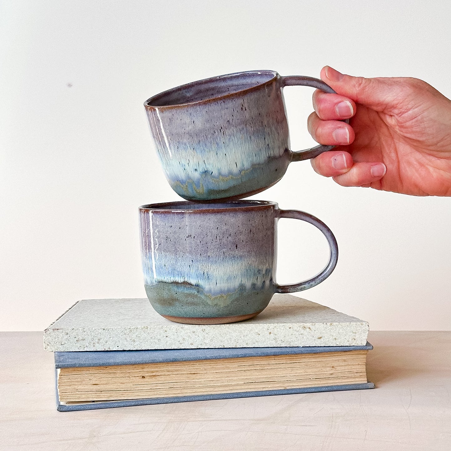 Two ceramic mugs stacked on a book with a hand holding one mug.