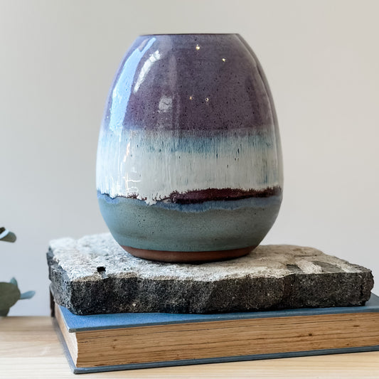 The Coastal Handmade Ceramic Round Vase sits on a tile and wooden table. The vase has a purple, white and blue glaze.