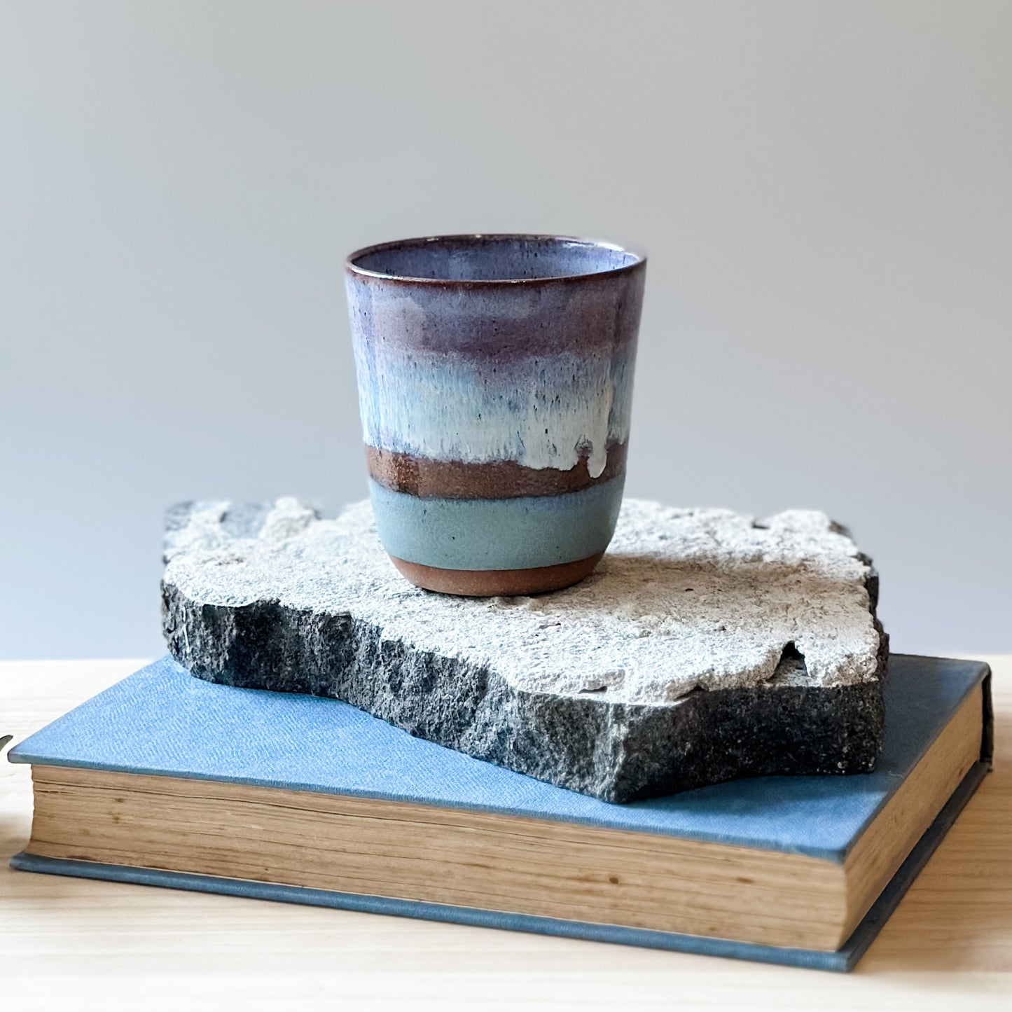 A handmade ceramic keepcup in a purple, blue and white glaze. The keep-cup sits on a tile and a wooden table.