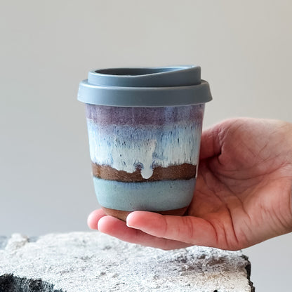 A handmade ceramic keepcup in a purple, blue and white glaze. The keep-cup is held above a tile.
