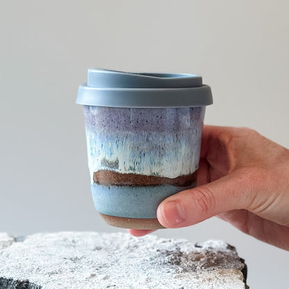 A handmade ceramic keepcup in a purple, blue and white glaze. The keep-cup is held above a tile.