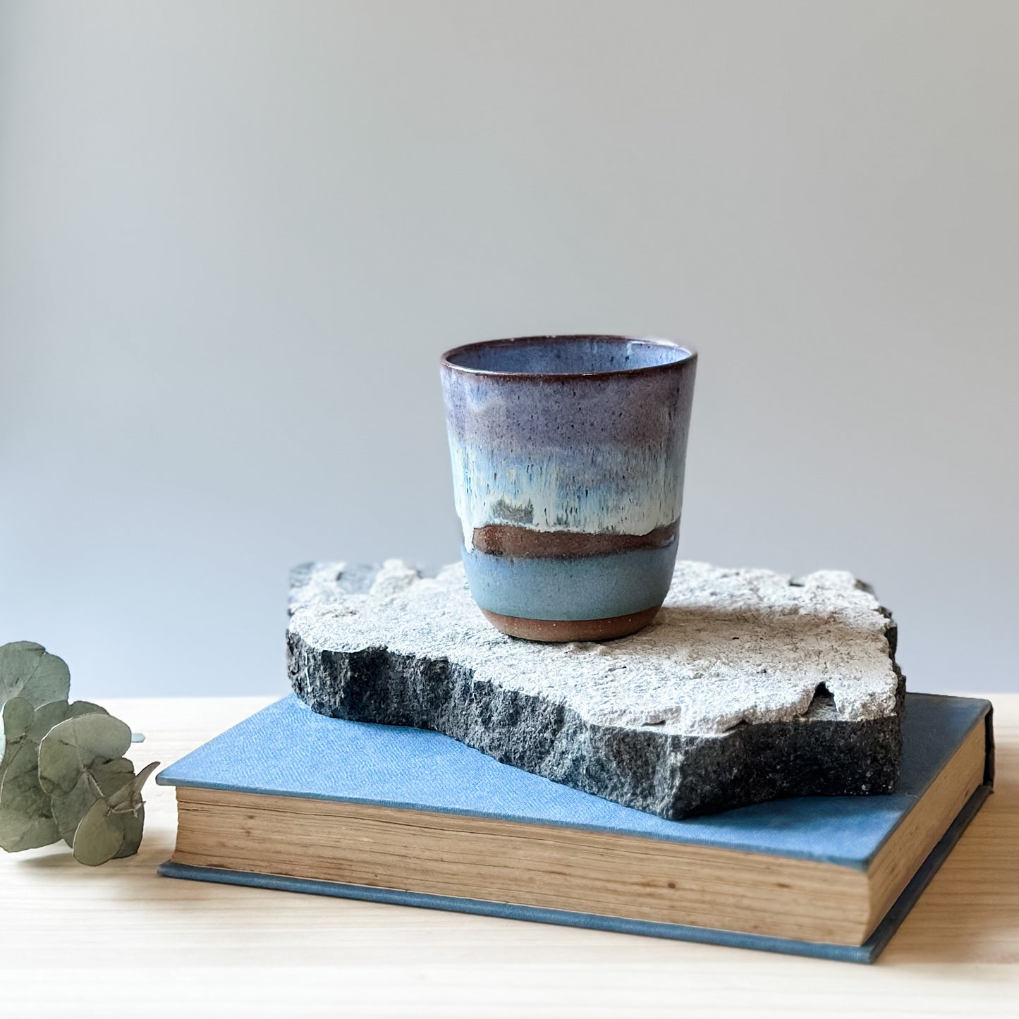 A handmade ceramic keepcup in a purple, blue and white glaze. The keep-cup sits on a tile and a wooden table.