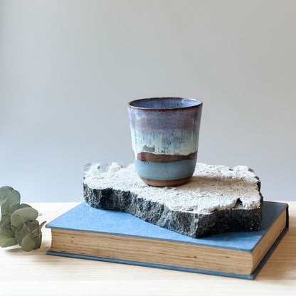 A handmade ceramic keepcup in a purple, blue and white glaze. The keep-cup sits on a tile and a wooden table.