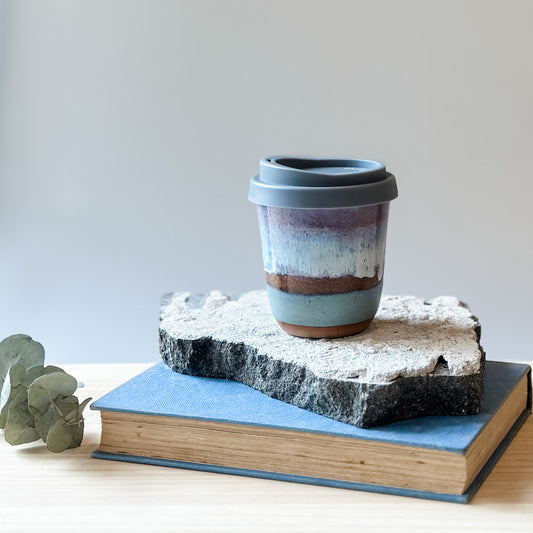 A handmade ceramic keepcup in a purple, blue and white glaze with a grey lid. The keep-cup sits on a tile and a wooden table.