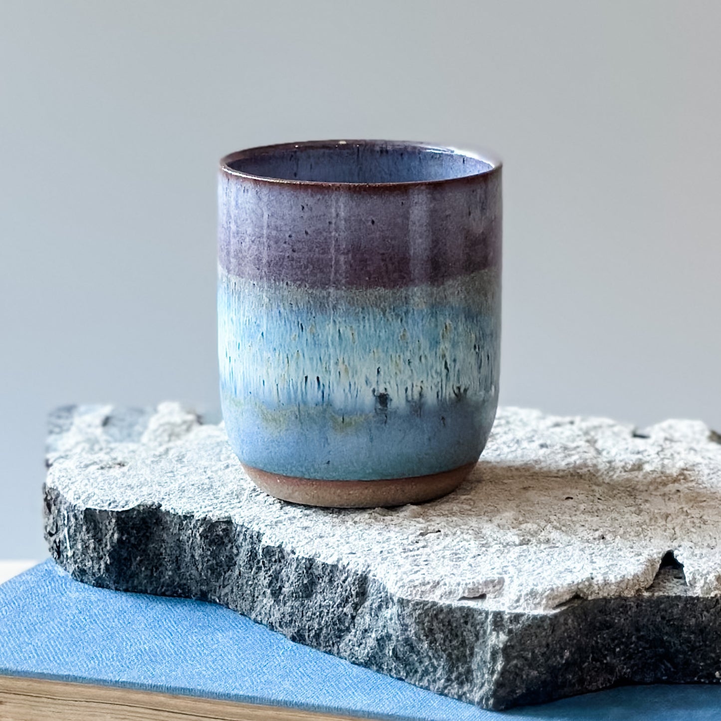 A close-up view of a coastal handmade ceramic tumbler glazed lavender, white and blue. The tumbler sits on a tile and a book.