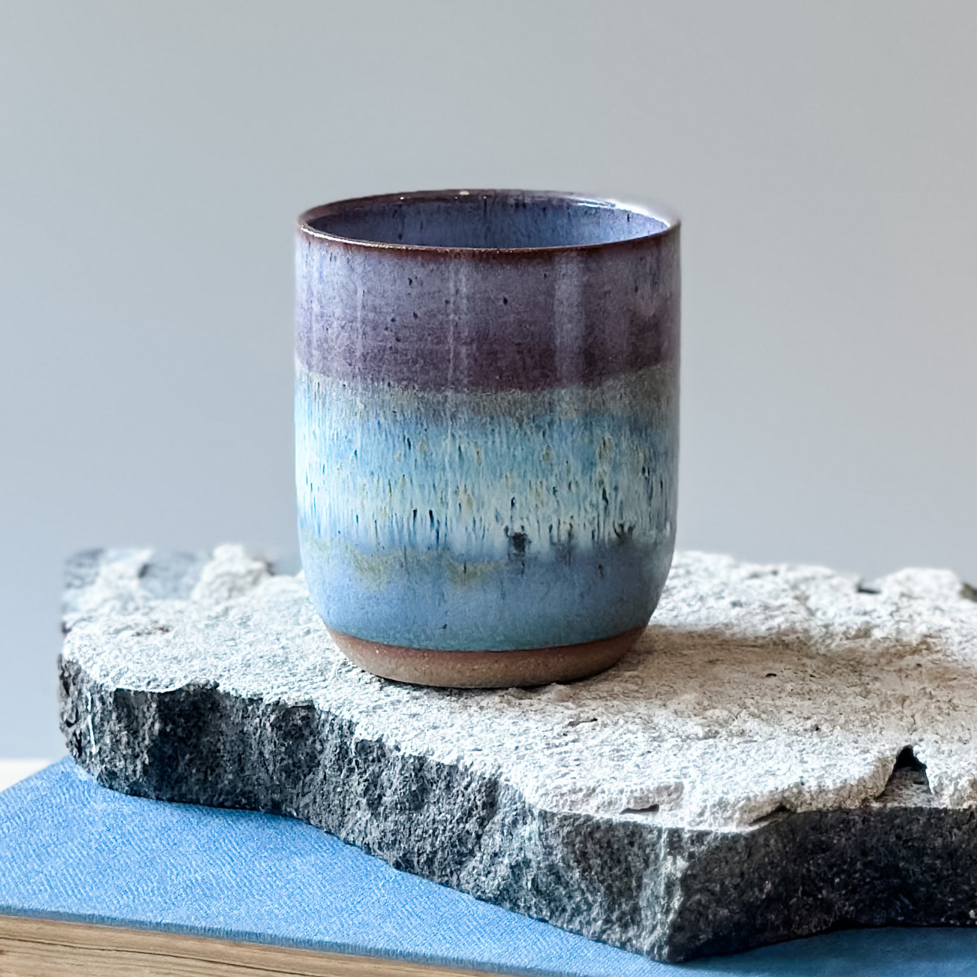 A close-up view of a coastal handmade ceramic tumbler glazed lavender, white and blue. The tumbler sits on a tile and a book.