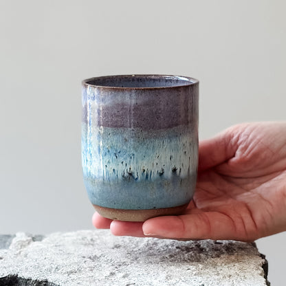 A coastal handmade ceramic tumbler glazed lavender, white and blue. The tumbler is held above a tile.