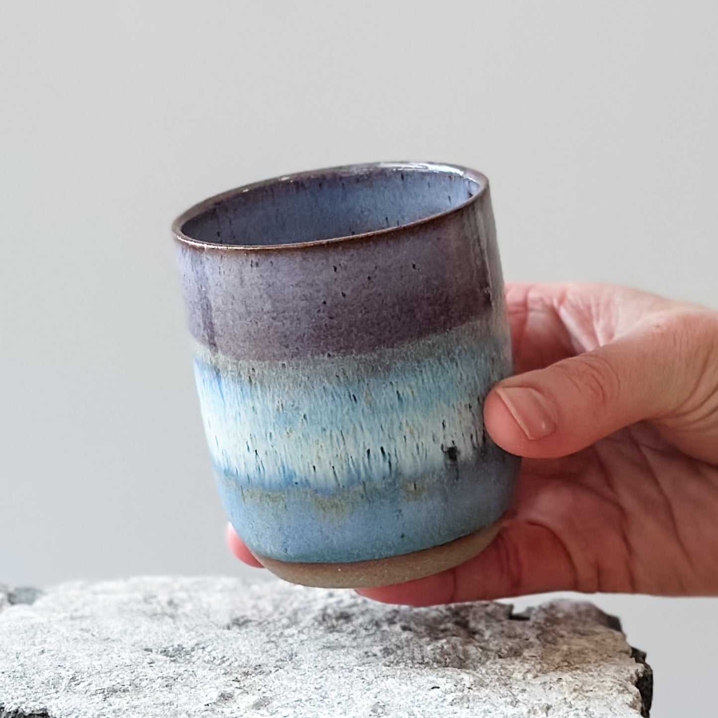 A coastal handmade ceramic tumbler glazed lavender, white and blue. The tumbler is held above a tile.