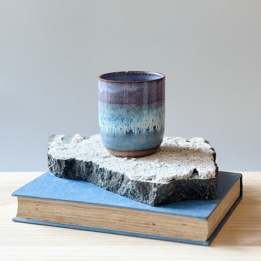 A coastal handmade ceramic tumbler glazed lavender, white and blue. The tumbler sits on a tile, a book, and a wooden table.
