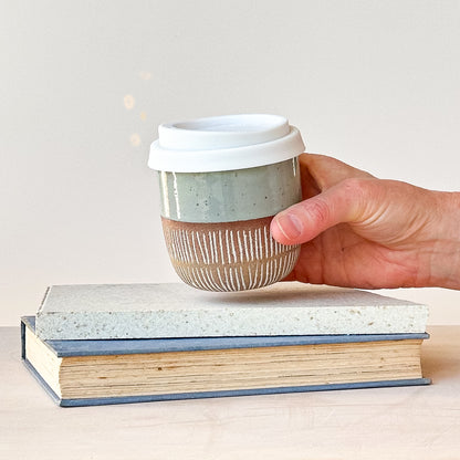 Hand holding a ceramic coffee cup with a white lid over two stacked books on a light background