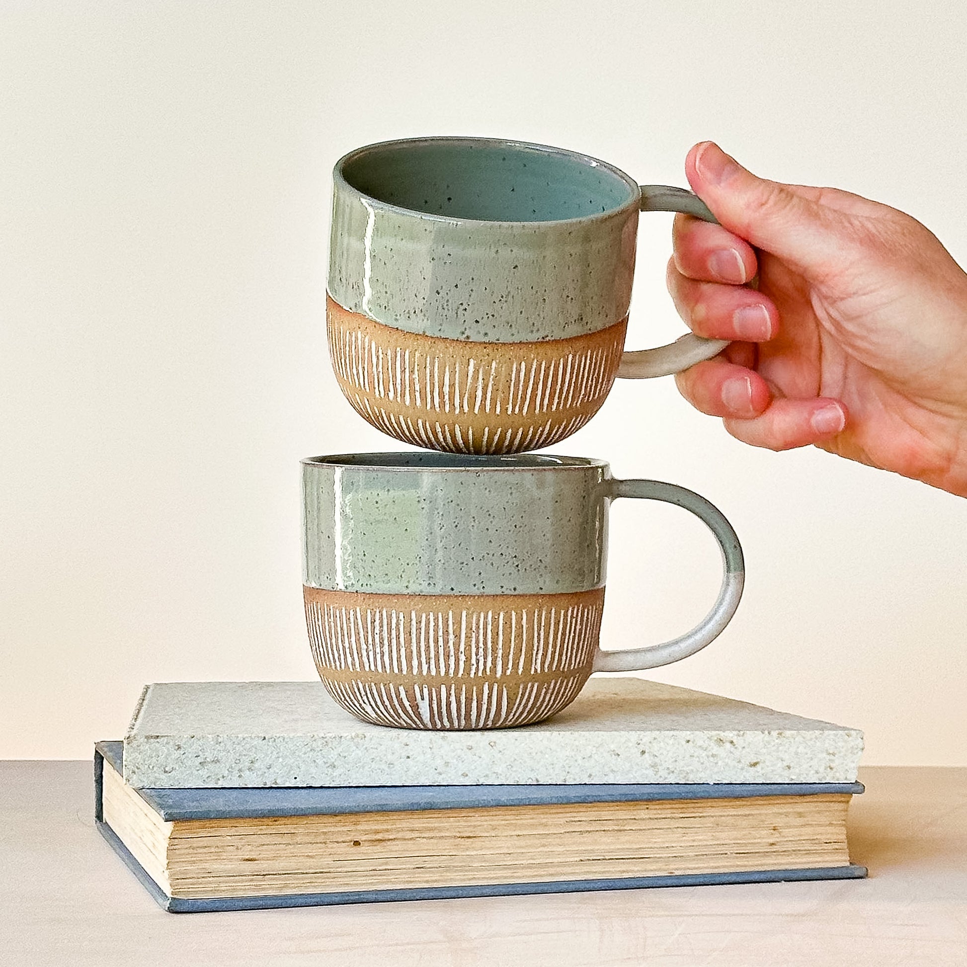 Two stacked ceramic mugs with a hand lifting one of them, on a light background.
