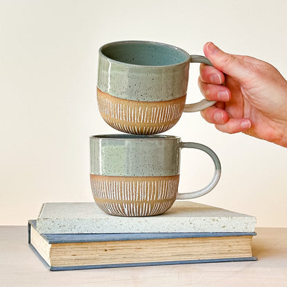 Two stacked ceramic mugs with a hand lifting one of them, on a light background.