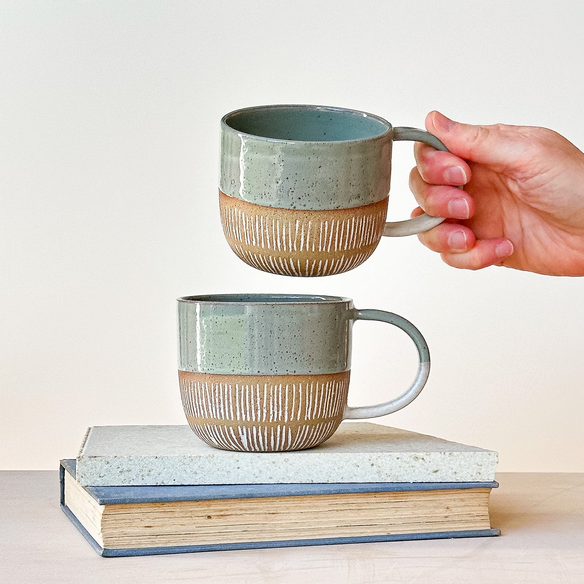 Two ceramic mugs with striped design, one held above the other, on a stack of books against a light background.