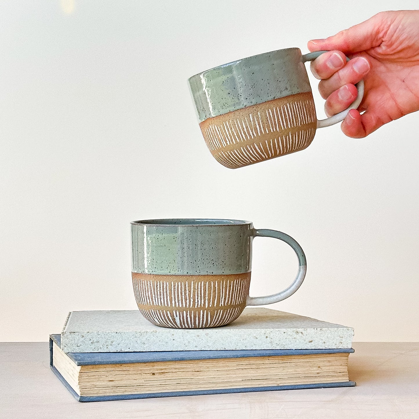 Person holding a ceramic mug above two stacked books on a light surface.