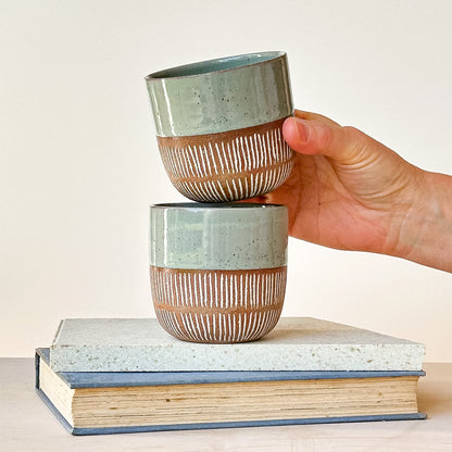 Hand holding a ceramic cup over another cup on top of books with a plain background