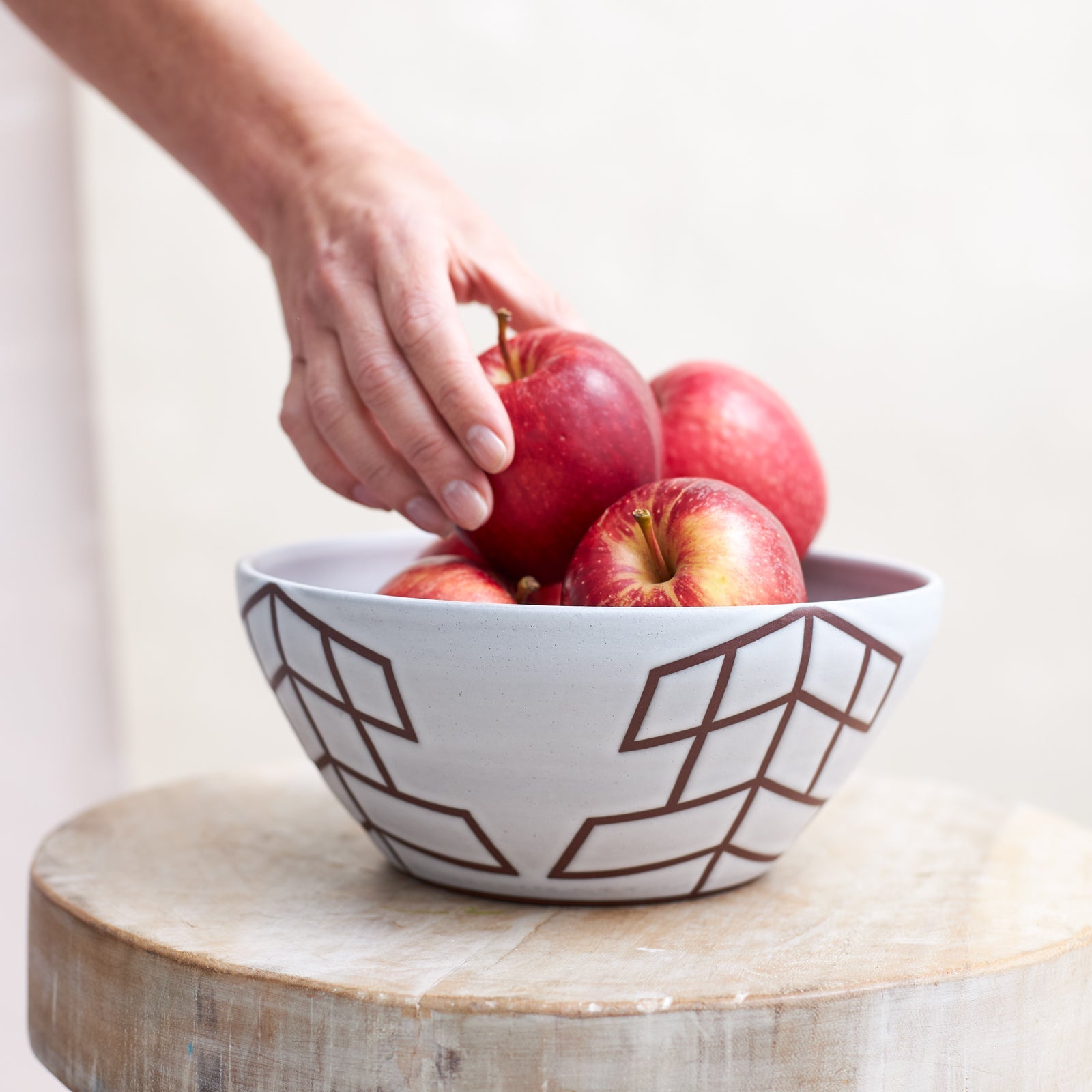 A front view of the 'Geometric Handmade Ceramic Bowl' with a white glaze and mahogany clay. Apples are placed by hand into the ceramic bowl. The handmade ceramic bowl sits on a wooden stool.