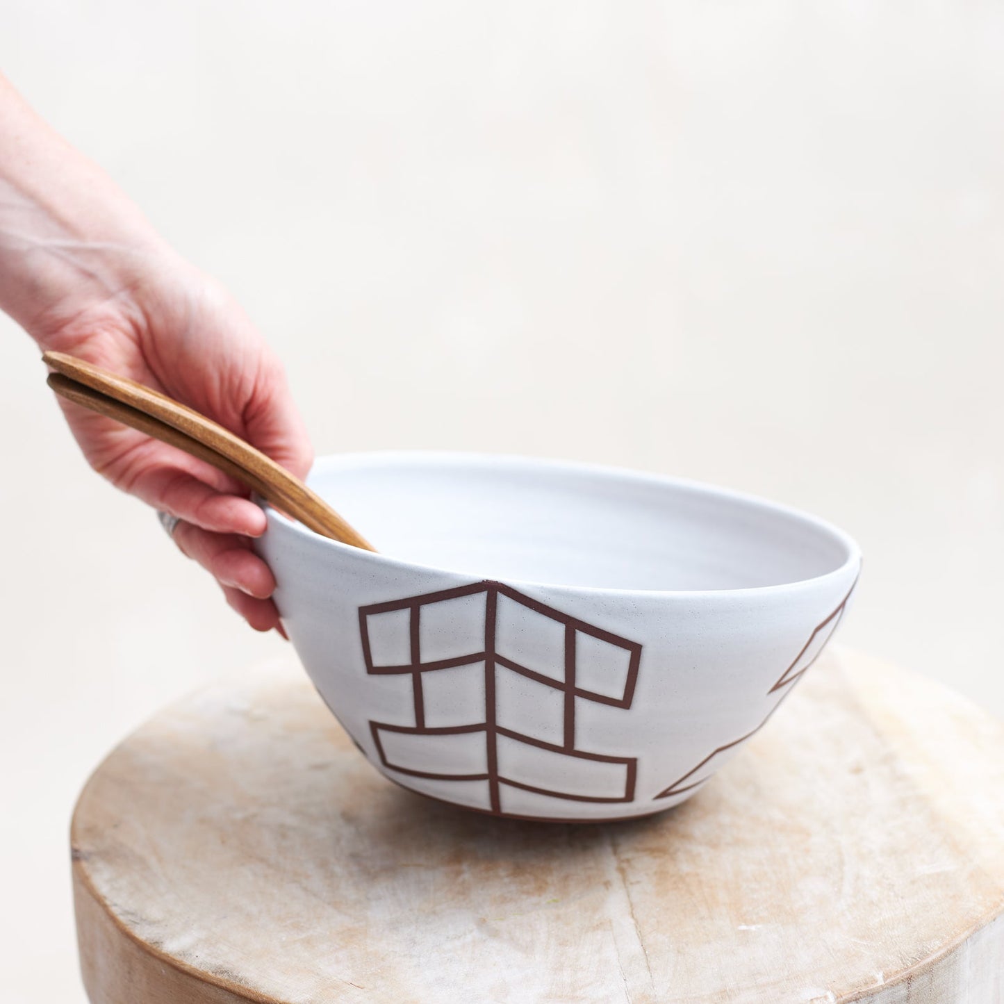  A front view of the 'Geometric Handmade Ceramic Bowl' with a white glaze and mahogany clay. Wooden salad servers are displayed in the ceramic bowl. The handmade ceramic bowl is being lifted by hand from a wooden stool.