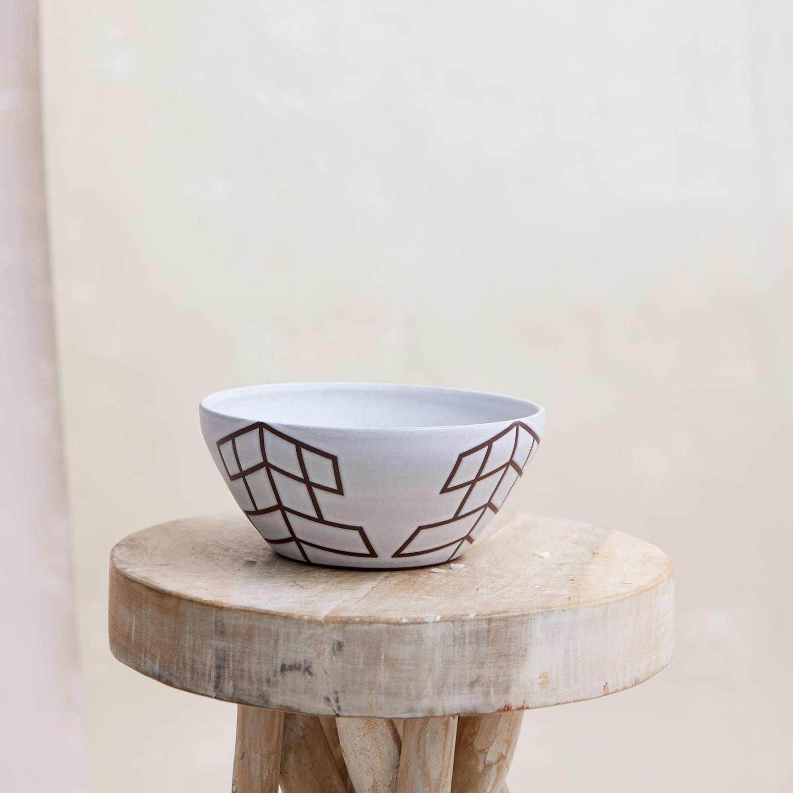 A front view of the 'Geometric Handmade Ceramic Bowl' with a white glaze and mahogany clay. The handmade ceramic bowl sits on a wooden stool in a coastal-styled setting.