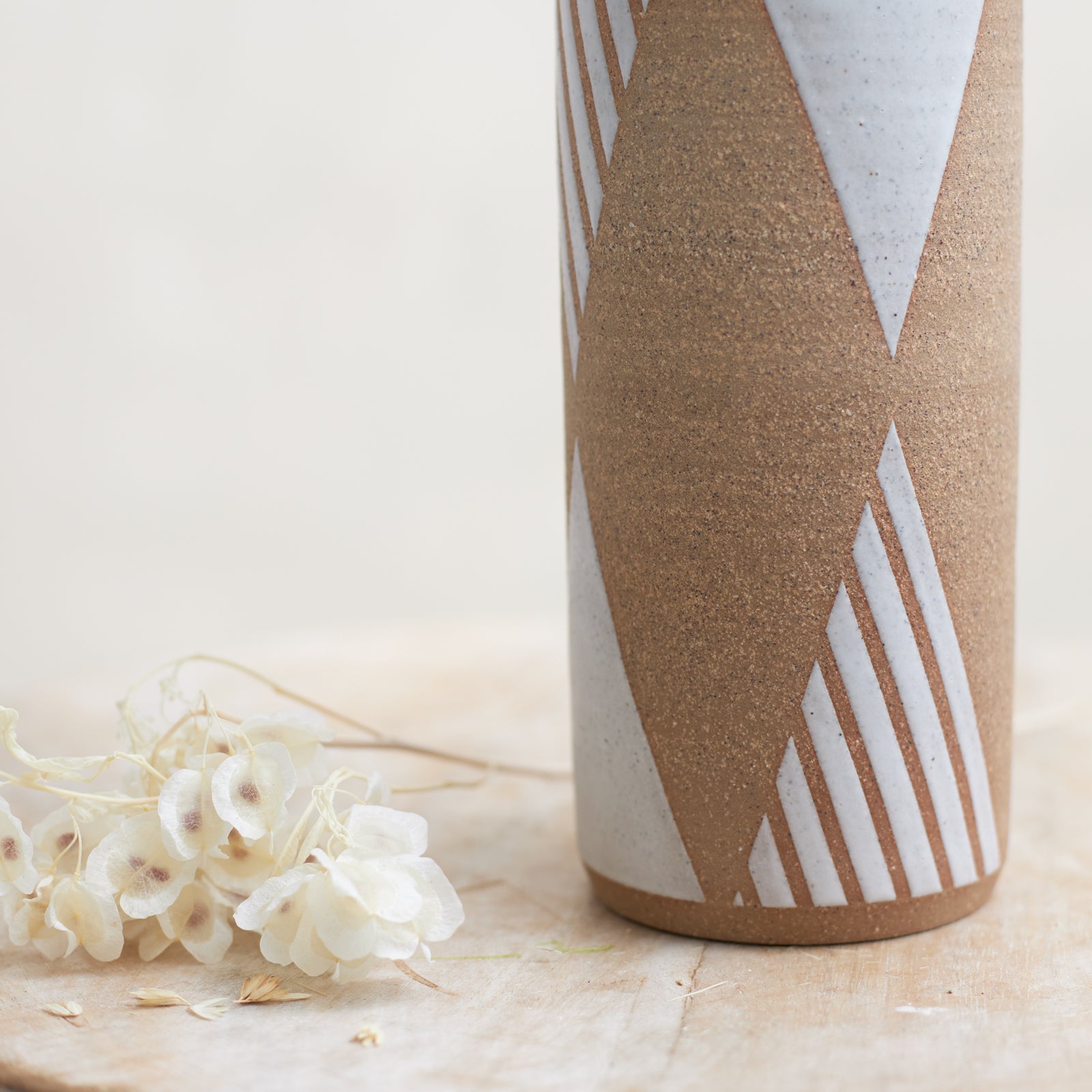 A close-up view of the geometric design on the 'Geometric Cylindrical Handmade Ceramic Vase' in natural and white glaze. The handmade vase sits on a wooden stool with flowers placed beside it. The vase sits in a coastal-styled setting.