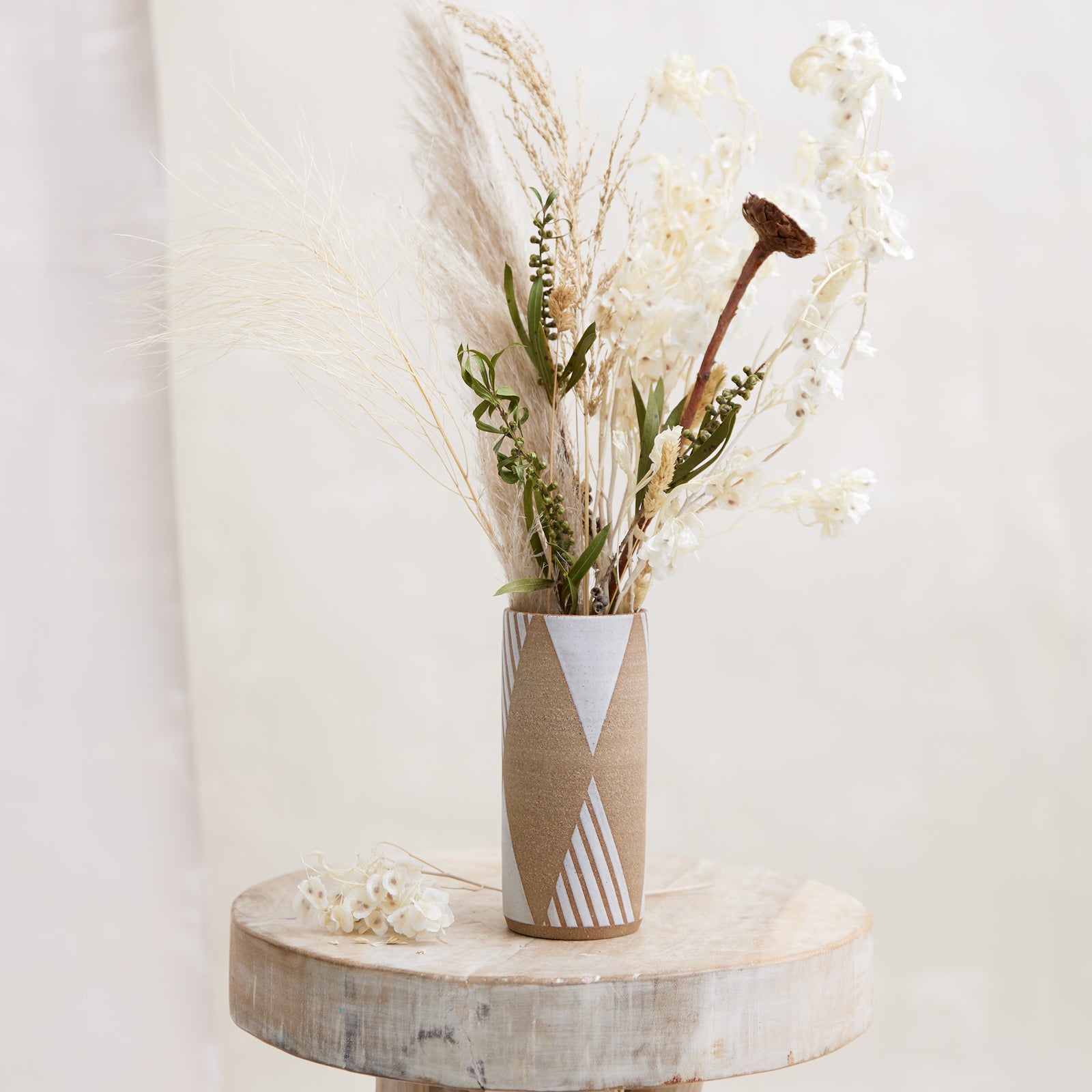 A front view of the 'Geometric Cylindrical Handmade Ceramic Vase' in natural and white glaze. The handmade vase sits on a wooden stool with flowers placed beside it. The vase sits in a coastal-styled setting. The handmade ceramic vase displays a dried flower arrangement.