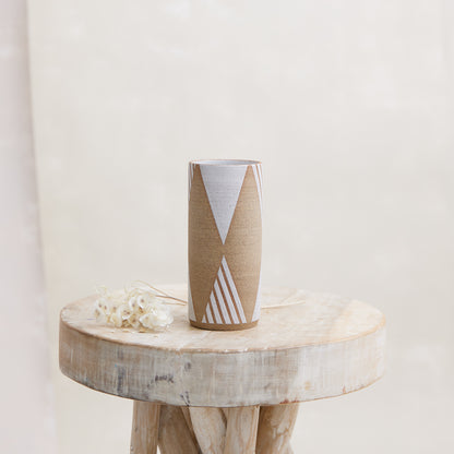 A front view of the 'Geometric Cylindrical Handmade Ceramic Vase' in natural and white glaze. The handmade vase sits on a wooden stool with flowers placed beside it. The vase sits in a coastal-styled setting.