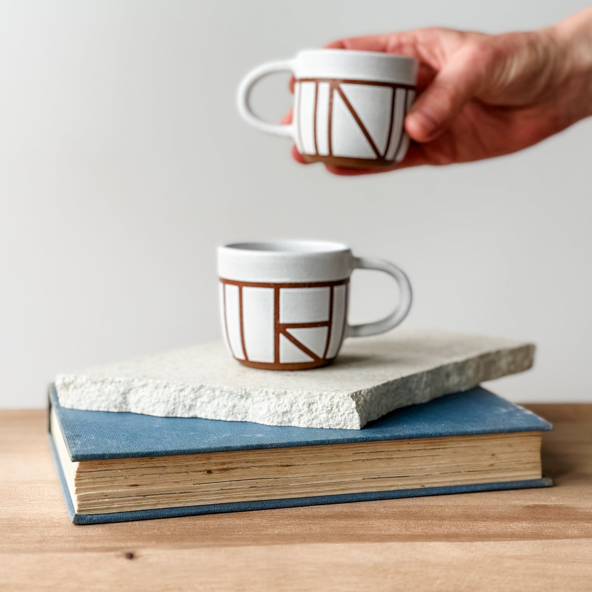 Two ceramic mugs with geometric patterns on a stack of books, one mug being held by a hand.