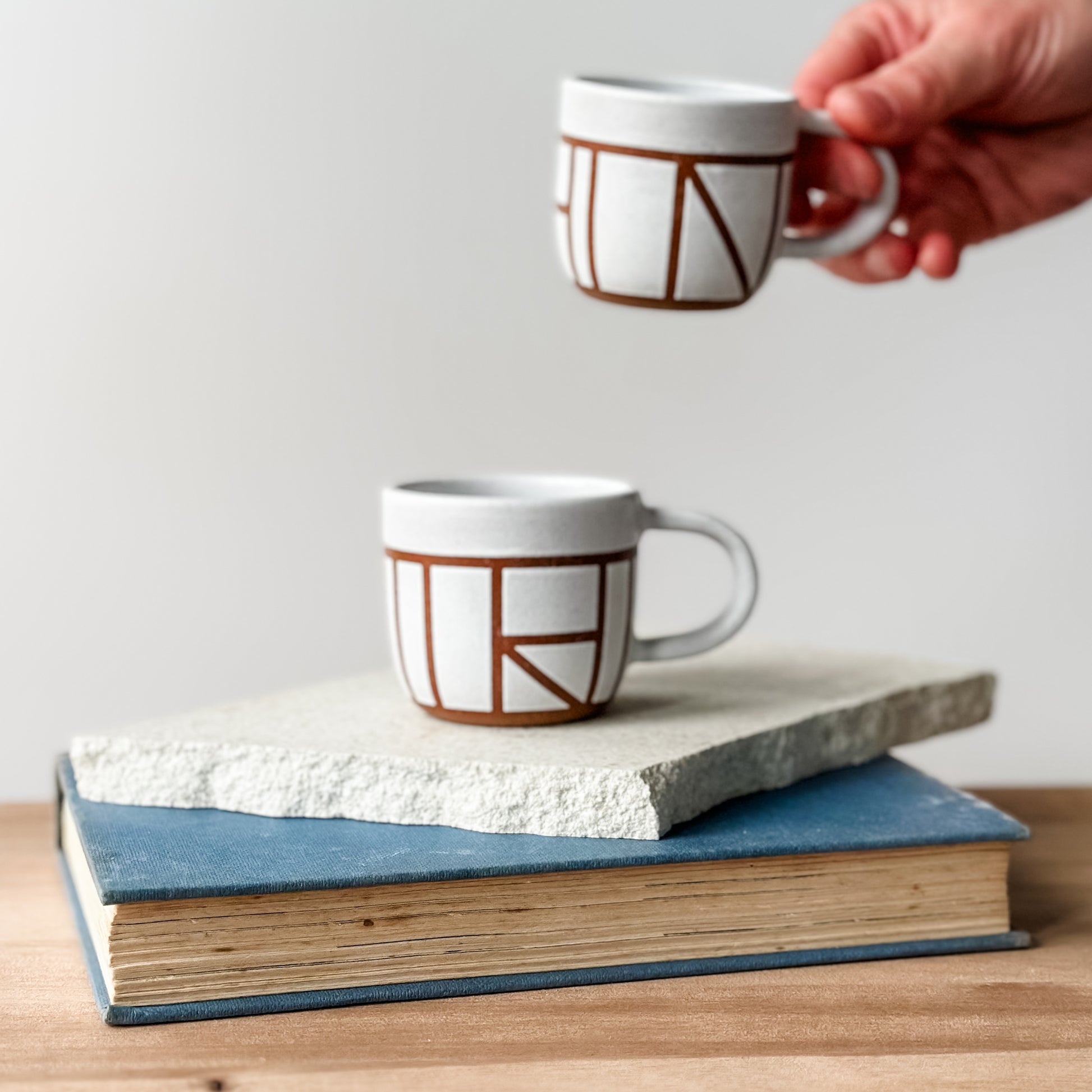 Two white mugs with brown geometric patterns on a stack of books, one held by a hand.