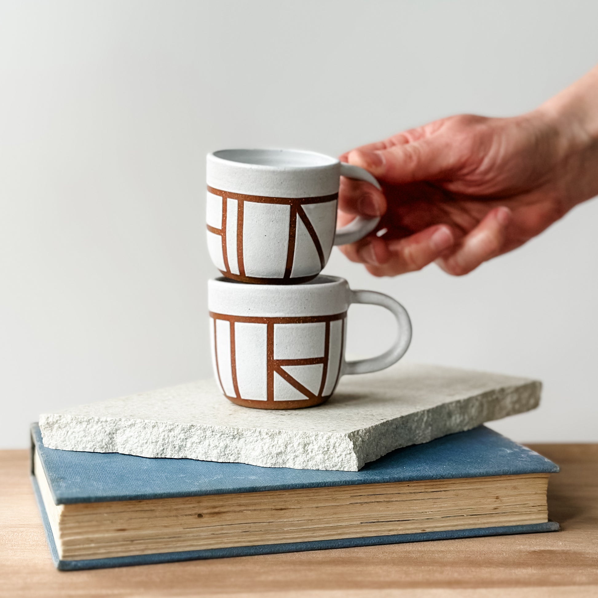Person holding a geometric-patterned mug over two stacked books with another mug on top.