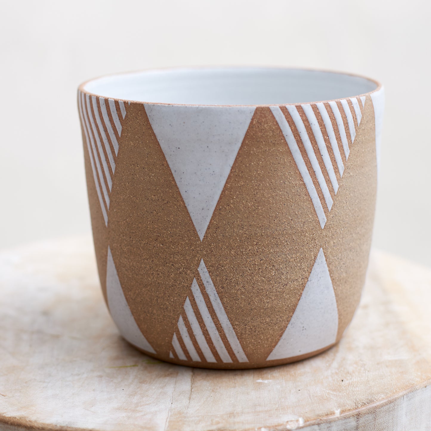 A close-up view of the Geometric Handmade Ceramic Planter Pot in a white glaze. The handmade planter sits atop a wooden stool in a coastal-styled setting.