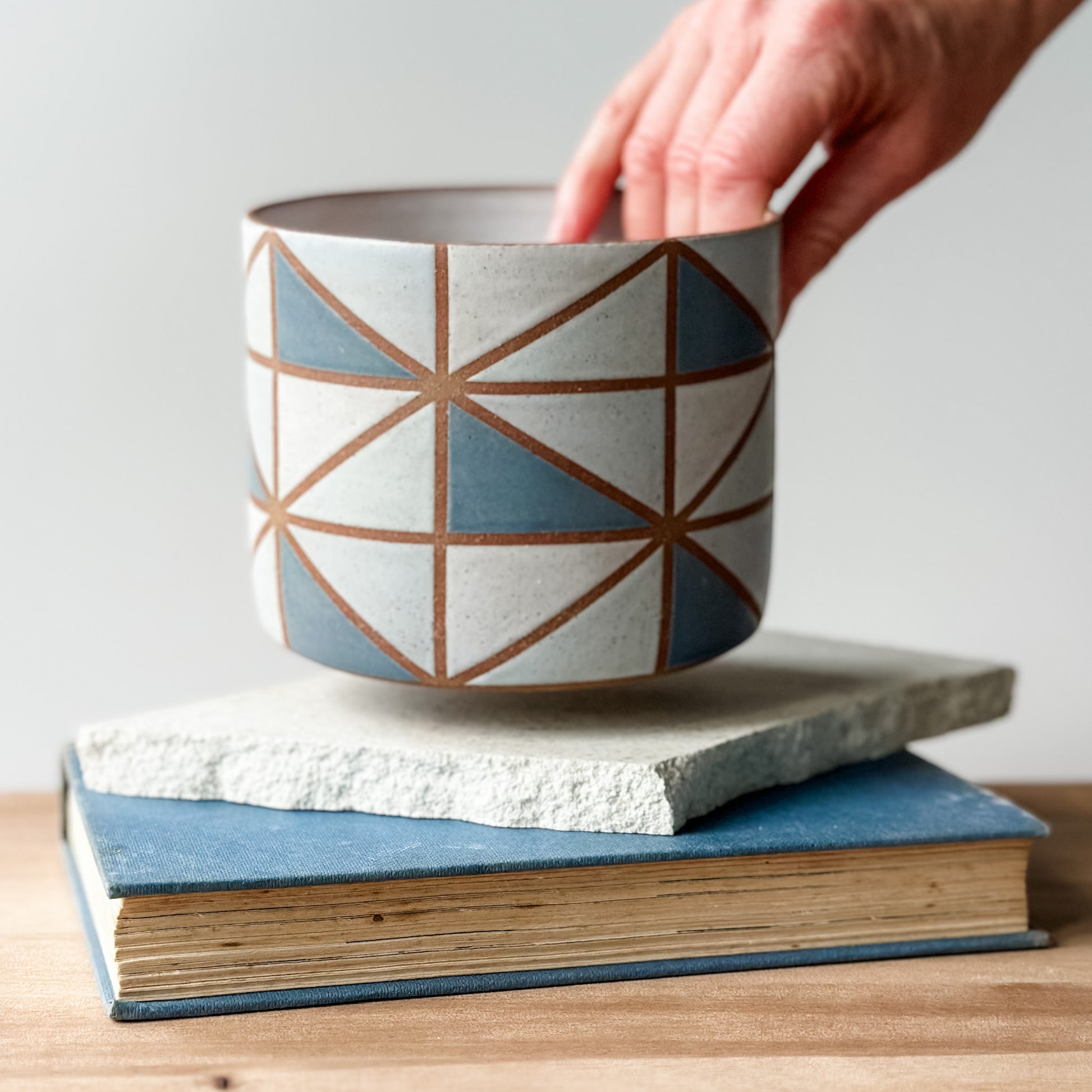 Ceramic pot with geometric design being held over a stack of books on a wooden surface.
