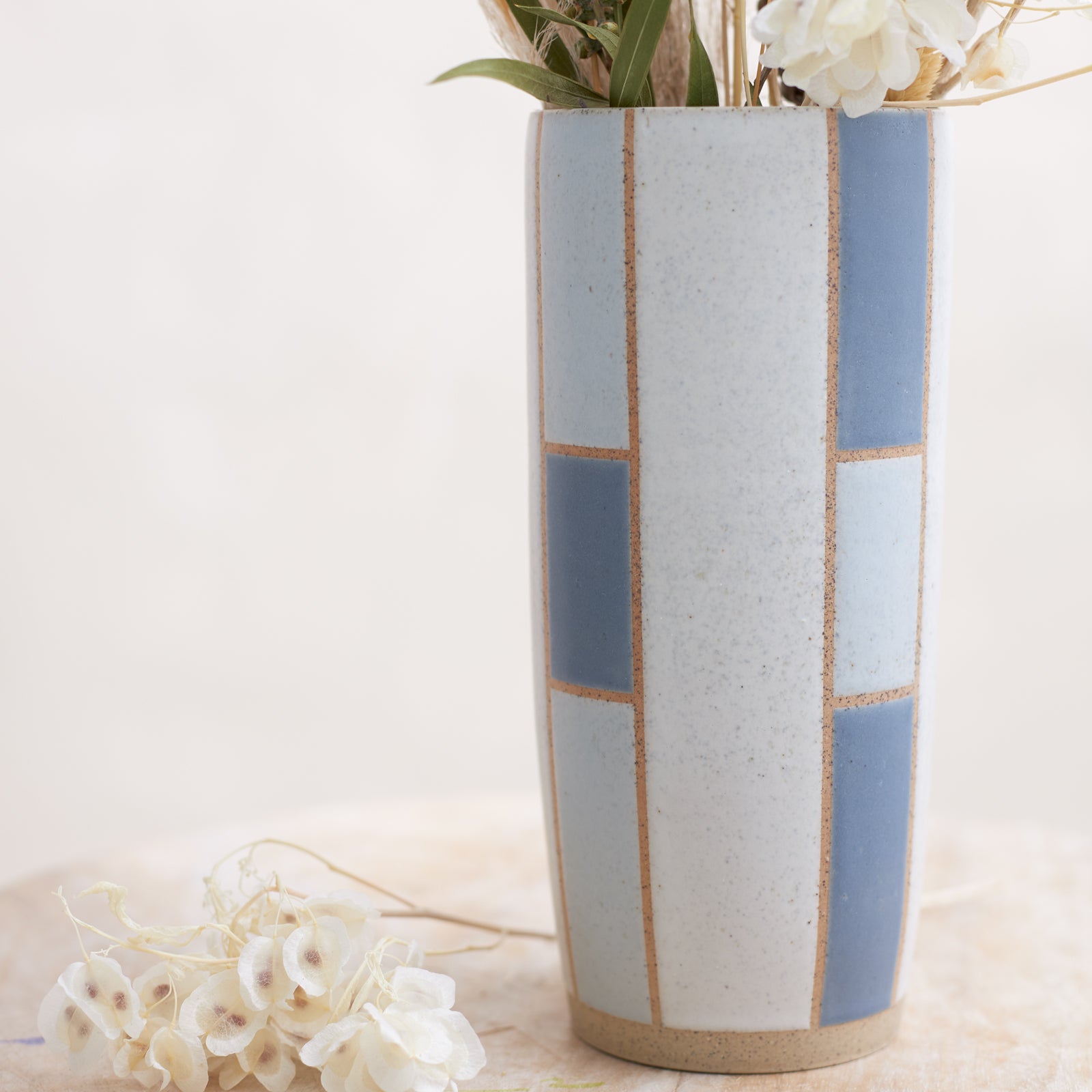 A close-up view of the Geometric Tapered Handmade Ceramic Vase in dark blue and grey glaze. The ceramic vase sits on a wooden stool holding dried flowers. The handmade vase is in a coastal-styled setting.