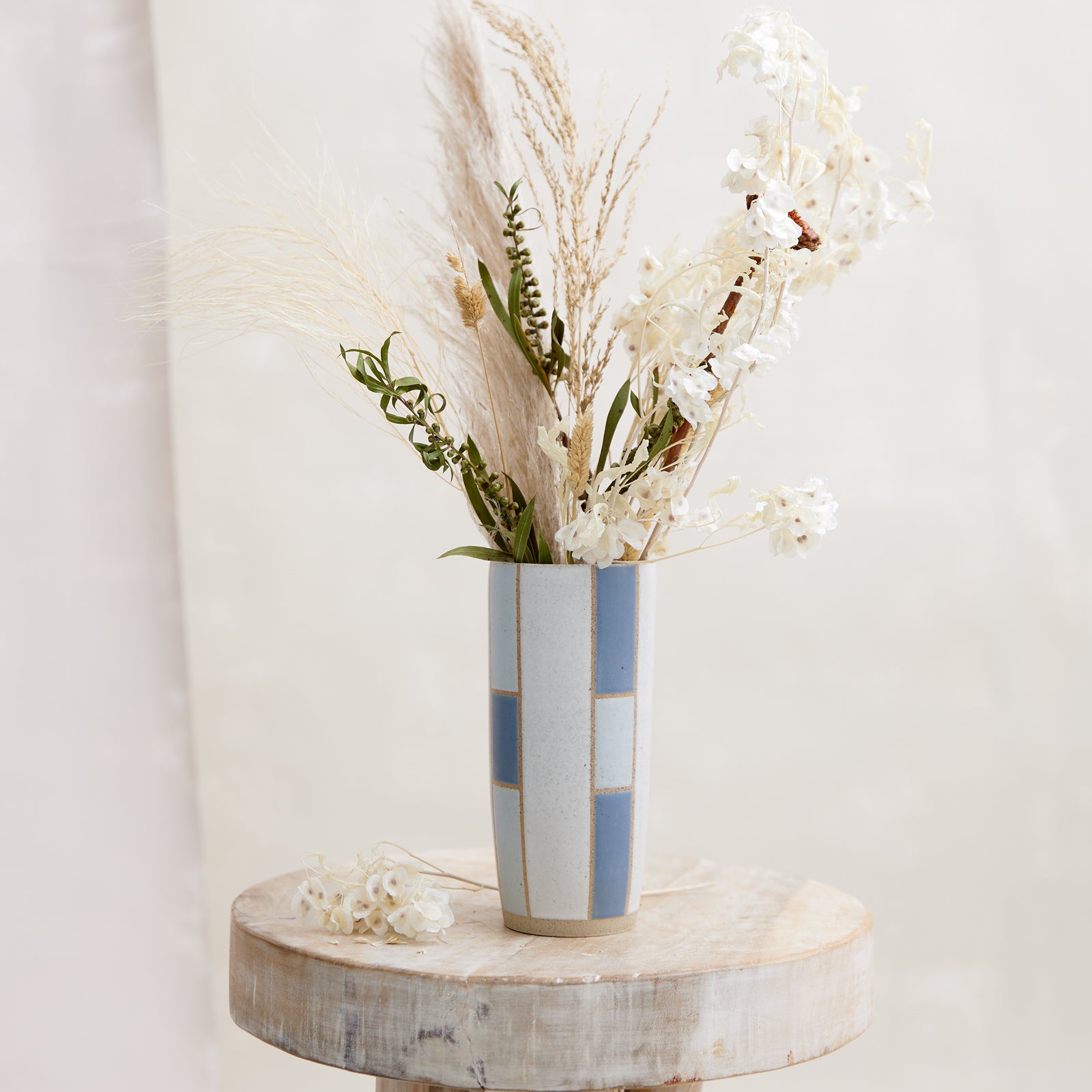 A front view of the Geometric Tapered Handmade Ceramic Vase in dark blue and grey glaze. The ceramic vase sits on a wooden stool holding dried flowers. The handmade vase is in a coastal-styled setting.