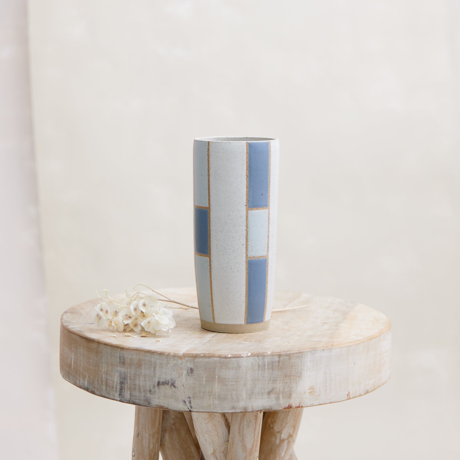 A front view of the Geometric Tapered Handmade Ceramic Vase in dark blue and grey glaze. The vase sits on a wooden stool next to dried flowers. The handmade vase is in a coastal-styled setting.
