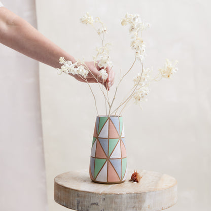 A front view of the Geometric Handmade Ceramic Teardrop Vase in pastel-coloured glazes. This handmade vase holds coastal dried flowers and sits on a wooden stool. A hand is arranging the dried flowers.